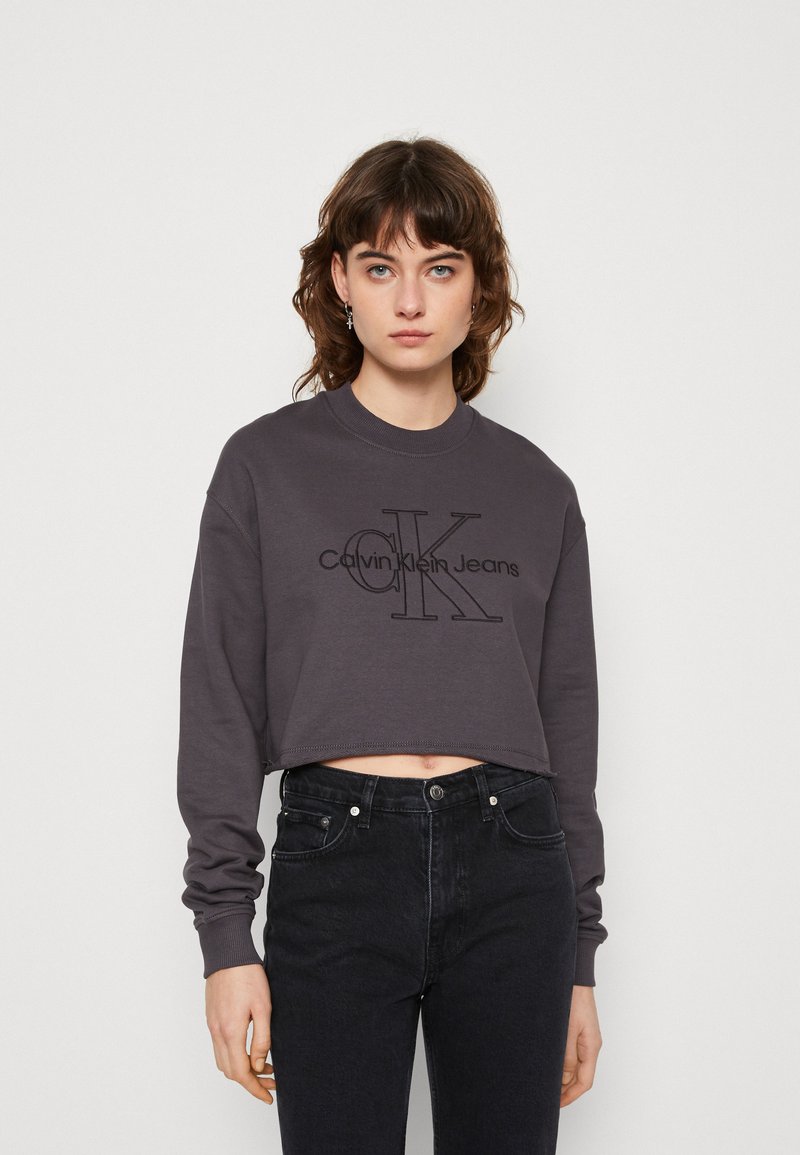 Kurtā grizā džemperis, izgatavots no kokvilnas, ar lielu melnu "Calvin Klein Jeans" logo krūšu rajonā un ribotiem piedokiem.