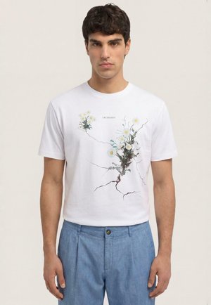 Mann trägt ein weißes T-Shirt mit einem floralen Design von Gänseblümchen, die durch Risse wachsen, kombiniert mit hellblauen Hosen.