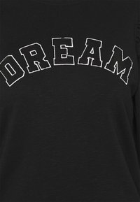 Camiseta negra con la palabra "DREAM" bordada en grandes letras en negrita y color blanco. La tela tiene un aspecto texturizado y ligeramente rayado.