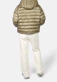 Veste verte olive gonflée avec capuche, présentant des panneaux cousus ; associée à un pantalon blanc et des chaussures grises. La veste a une texture brillante.