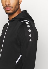 Zwarte sportieve hoodie met een rits aan de voorkant, textuurstof, witte accenten en ronde reflecterende details op de schouder. Glad afwerking.