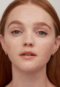 Retrato de una persona con piel clara y pecas, cabello castaño claro, maquillaje suave y pendientes de aro, que muestra una textura suave y rasgos naturales.
