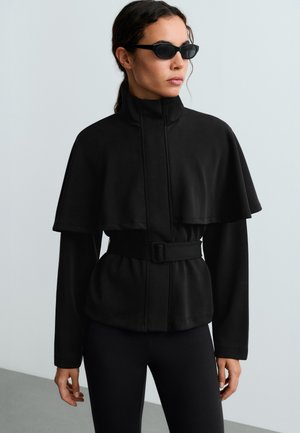 Femme portant une veste noire ceinturée à col montant avec un détail cape et des leggings noirs, regardant sur le côté, avec des lunettes de soleil foncées.