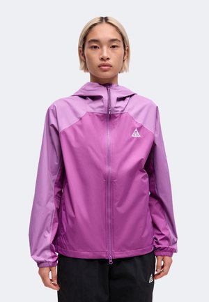 Personne portant une veste à capuche zippée bicolore violette et un pantalon noir avec un logo sur la poitrine gauche et la cuisse gauche.