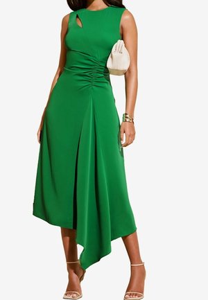 Robe midi verte sans manches avec ourlet asymétrique, fronces latérales, associée à des talons beiges, un bracelet manchette doré et un petit sac à main beige.