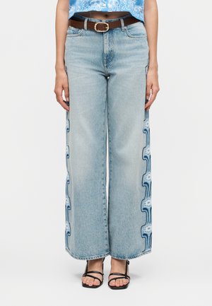 Lyseblå jeans med brede ben med blå og hvide blomsterbroderier langs sidesømme, kombineret med et brunt bælte og sorte sandaler med remme.