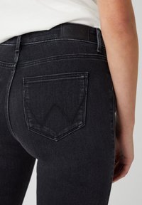 Jeans de mezclilla negra con un diseño ajustado, que presentan un bolsillo con una "W" bordada y una parche de cuero en la cintura.