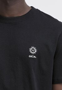 Tricou din bumbac negru cu guler rotund, mâneci scurte și un soare brodat alb și textul "calm." în zona stângă a pieptului.
