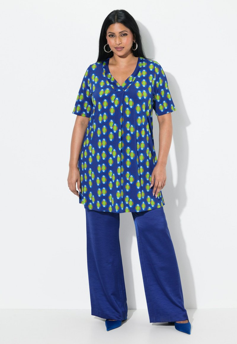 Blaue Tunika mit grünem und gelbem blattähnlichem Muster, kurzen Ärmeln und V-Ausschnitt. Kombiniert mit weiten blauen Hosen und blauen High Heels.
