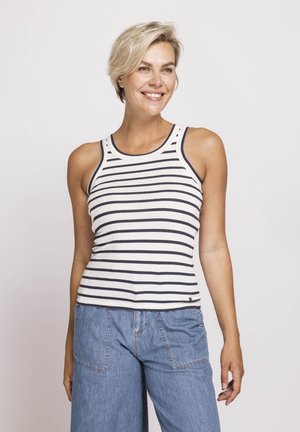 Smiling woman with short blonde hair wearing a white tank top with black stripes and light blue denim pants against a plain background.