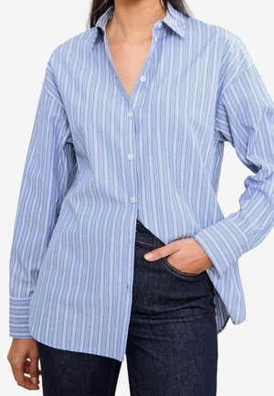 Mujer con camisa de manga larga azul claro con rayas blancas, combinada con vaqueros azul oscuro, una mano en el bolsillo.