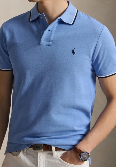 Ανοιχτό μπλε πόλο πουκάμισο με υφή στο ύφασμα, γιακά σε αντίθεση και μικρό σκουρόχρωμο λογότυπο στο στήθος. Φορεμένο με λευκά σορτς και καφέ ζώνη.