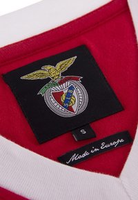 Rode stof met een witte kraag, voorzien van een zwart geborduurd embleem met een gouden adelaar en een voetbalinsigne. Label leest "Gemaakt in Europa."