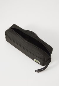 Trousse noire en tissu lisse, à fermeture éclair, avec un logo de crocodile vert. Elle présente un design mince et rectangulaire.