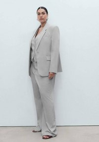 Svetle šedý oblek s jemným proužkem, obsahující jednoradový blazer, odpovídající vestu a široké kalhoty, doplněný červenohnědými sandály na pásky.