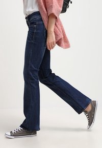 Donkerblauwe rechte jeans met een lichte vervaging, gecombineerd met grijze canvas sneakers met witte zolen en veters, en een zachte roze cardigan.