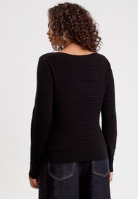 Pull noir à texture avec des manches longues et un col rond. Il présente un ourlet et des poignets côtelés, affichant un motif uniforme.