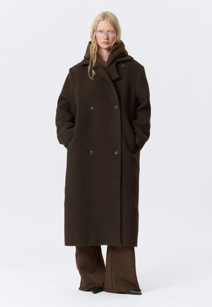 Manteau double boutonnage en laine marron avec un grand col, des poches latérales et une coupe décontractée, porté sur un ensemble marron assorti.