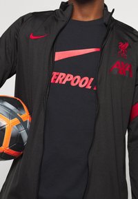 Sort zip-up jakke med røde detaljer, logo og branding. Underneath bærer en mørk t-shirt et stort rødt Nike og "LIVERPOOL" grafik. Holder en fodbold.