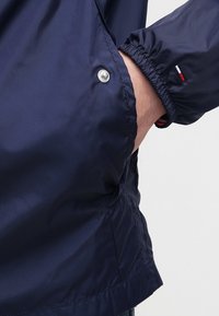 Námořnická modrá bunda s hladkým, lesklým povrchem. Obsahuje elastické manžety, detail na patky a tri-kulorový akcent na rukávu.