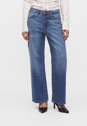 Personne portant un jean bleu droit, une chemise blanche à manches longues et des chaussures noires à talons pointus, debout devant un fond uni.