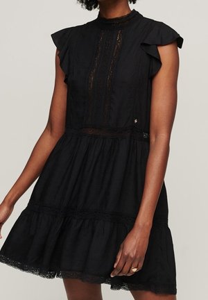 Femme portant une robe noire sans manches avec des détails en dentelle, des épaules à volants et une jupe à étages, debout devant un fond uni.