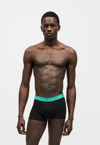 Άνδρας χωρίς πουκάμισο με μυώδη σωματότυπο που φοράει μαύρα boxers Tommy Hilfiger με πράσινη μέση, στέκεται μπροστά σε απλό ανοιχτό φόντο.