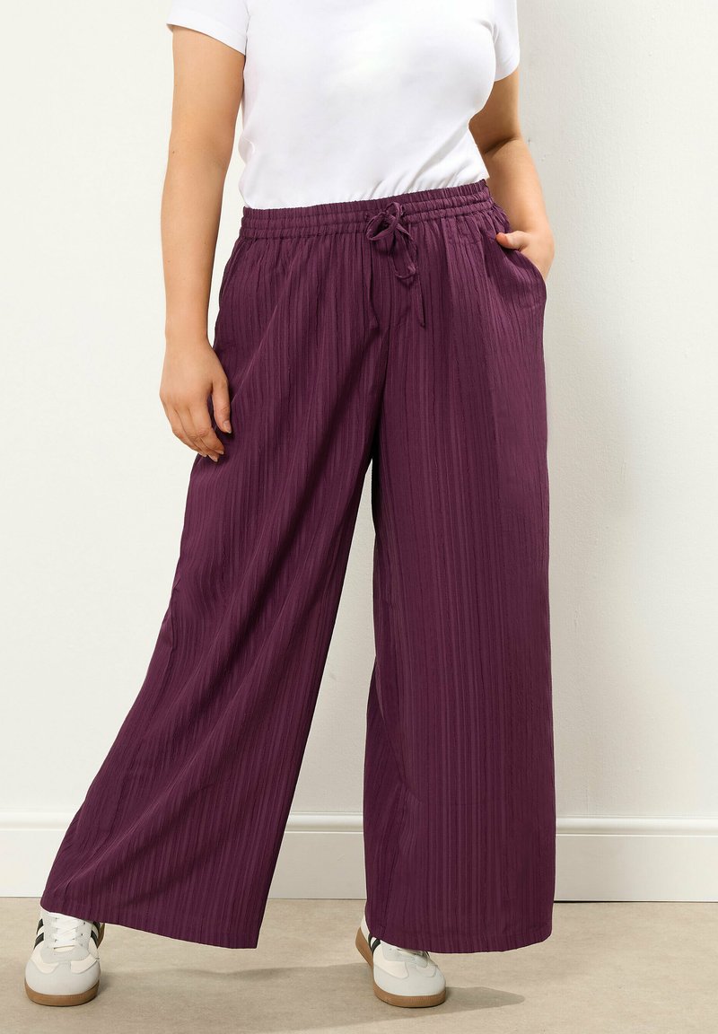 Szerokie spodnie w głębokim fioletowym kolorze z teksturowanym wykończeniem, wyposażone w elastyczny pas i sznurek. Stylizowane z białą bluzką i sneakersami.
