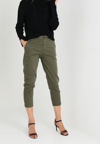 Pantaloni cropped verde oliva con un sottile motivo texturizzato, abbinati a un maglione nero in maglia e tacchi neri con strappi.