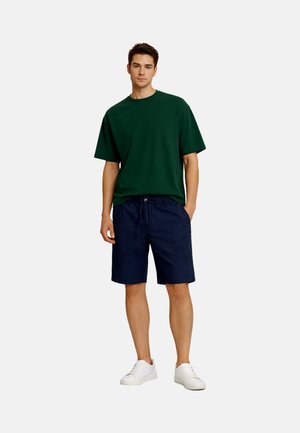 Junger Mann stehend, trägt ein dunkelgrünes übergroßes T-Shirt, marineblaue Shorts und weiße Turnschuhe vor einem schlichten hellen Hintergrund.