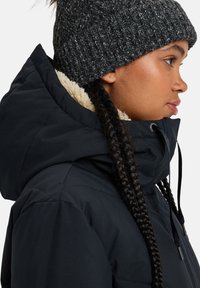 Chaqueta de plumas negra con un cuello alto y cordón, con un forro de forro polar beige. Usada con un gorro de punto gris texturizado.