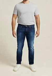 Homme portant un t-shirt gris clair à manches courtes, un jean bleu foncé et des baskets blanches, debout devant un fond beige uni.