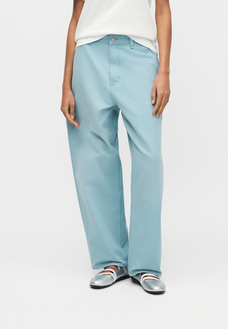 Personne portant un pantalon large bleu clair, une chemise blanche rentrée et des chaussures Mary Jane argentées métalliques, debout devant un fond uni.