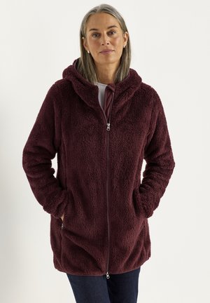 Frau mittleren Alters mit grauen Haaren, die eine dunkel bordeauxrote Fleecejacke mit Reißverschluss und Kapuze trägt und die Hände in den Taschen hat.