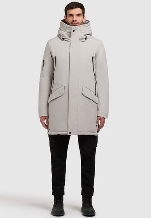 Lichtgrijze parka met een hoge kraag, ritssluiting aan de voorkant, zij- en borstzakken, en een gewatteerde textuur. Gecombineerd met zwarte cargobroek en laarzen.