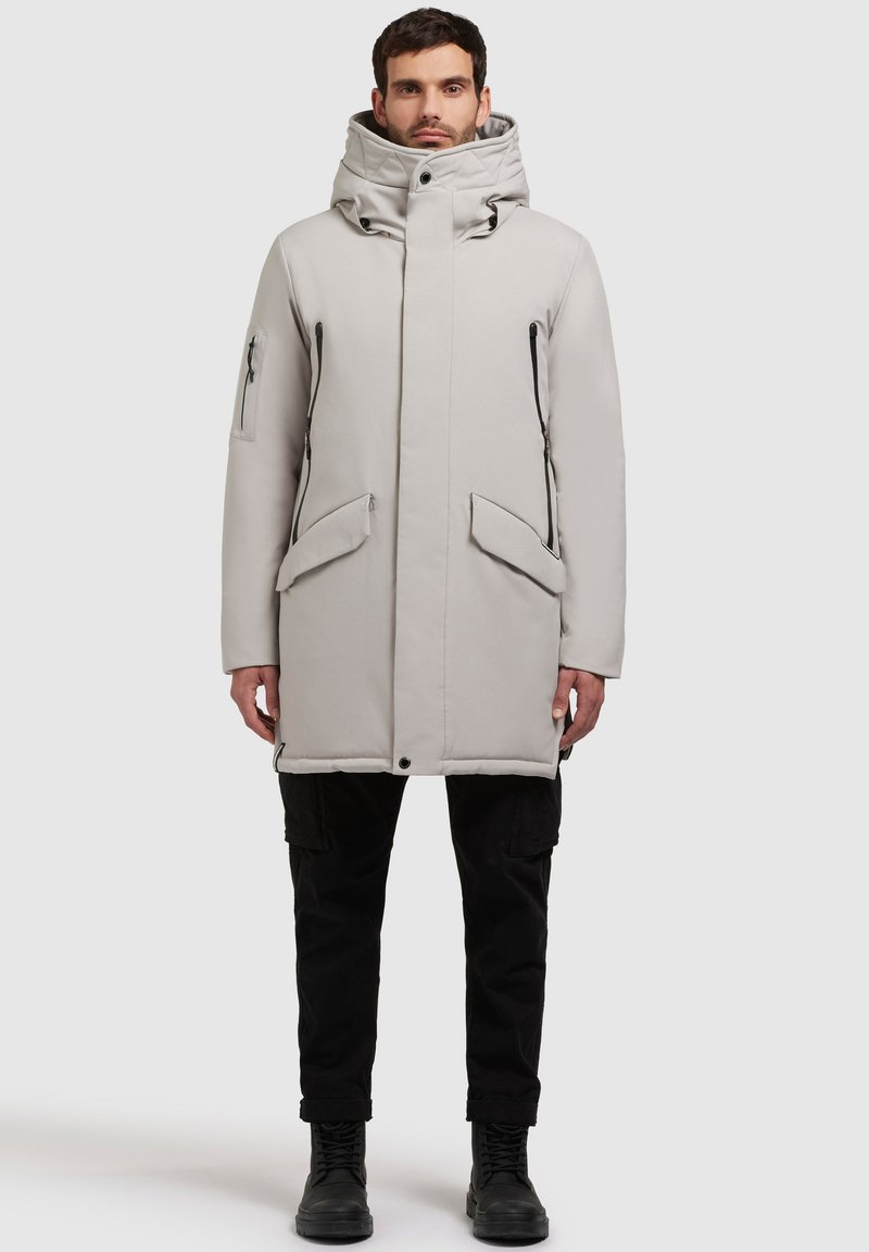 Parcă gri deschis cu guler înalt, închidere cu fermoar în față, buzunare laterale și pe piept și o textură capitonată. Asortat cu pantaloni cargo negri și cizme.