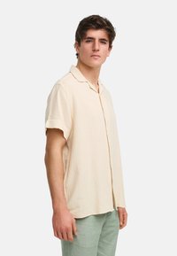 Camisa corto de manga corta color beige claro, hecha de tejido texturizado con un ajuste relajado, que presenta un cuello clásico y un sutil patrón de cuadrícula.