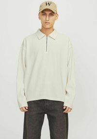 Suéter acanalado de color crema con un cuello con media cremallera, combinado con unos jeans oscuros. El modelo también lleva una gorra beige con una letra "W."