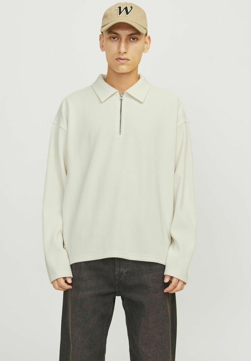 Suéter acanalado de color crema con un cuello con media cremallera, combinado con unos jeans oscuros. El modelo también lleva una gorra beige con una letra "W."