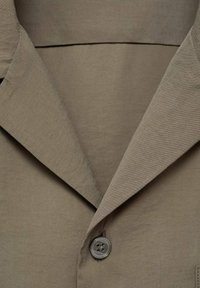 Khaki blazer met een ingezette kraag, een enkele knoopsluiting en een gladde stoftextuur. De knoop is rond met vier gaten.