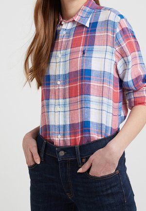 Kobieta ubrana w czerwoną, białą i niebieską koszulę w kratę, wsuniętą do ciemnoniebieskich dżinsów, z rękami w kieszeniach, z widocznymi długimi brązowymi włosami.