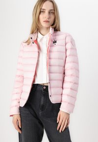 Giacca puffer di colore rosa chiaro con cuciture orizzontali, chiusura a bottoni e una patch con il logo sulla spalla. Abbinata a jeans neri.