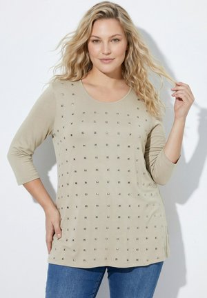 Blonde vrouw die een beige top met lange mouwen en vierkante studs draagt en een blauwe spijkerbroek, poserend tegen een witte achtergrond.