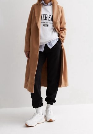 Person iført en camel-lang frakke, hvid grafisk sweatshirt, blåstripet skjorte, sorte joggebukser, hvide sokker og hvide sneakers, stående med hænderne i lommerne.