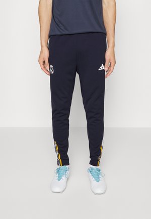 Pantalones de chándal azul marino con el logo del Real Madrid, marca Adidas, corte ajustado y acentos laterales a rayas amarillas. Combinados con zapatillas deportivas blancas.