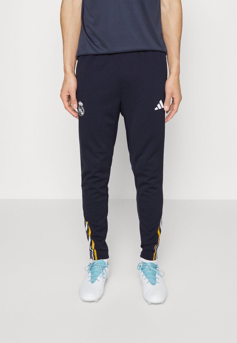 Pantalones de chándal azul marino con el logo del Real Madrid, marca Adidas, corte ajustado y acentos laterales a rayas amarillas. Combinados con zapatillas deportivas blancas.