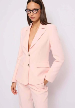 Donna che indossa un blazer rosa chiaro su misura e pantaloni abbinati, con occhiali neri a forma di occhio di gatto, che posa con una mano in tasca davanti a uno sfondo bianco.