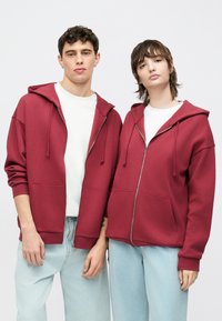 Bordó színű cipzáras kapucnis pulóverek elején zsebekkel, a kapucnin húzózsinórokkal. Fehér pólók fölött viselve, világoskék farmerral párosítva.