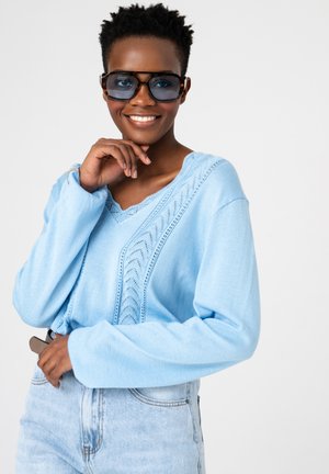 Uśmiechnięta osoba ubrana w niebieski, oversize'owy sweter, jasne dżinsy i duże przyciemniane okulary przeciwsłoneczne, pozująca z jedną ręką przy twarzy, a drugą na biodrze.