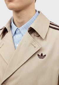 Άνδρας φοράει ένα μπεζ παλτό τύπου trench από την Adidas πάνω από ένα μπλε και άσπρο ριγέ πουκάμισο με κουμπιά, εμφανίζοντας το γιακά του παλτού και το λογότυπο στο στήθος.